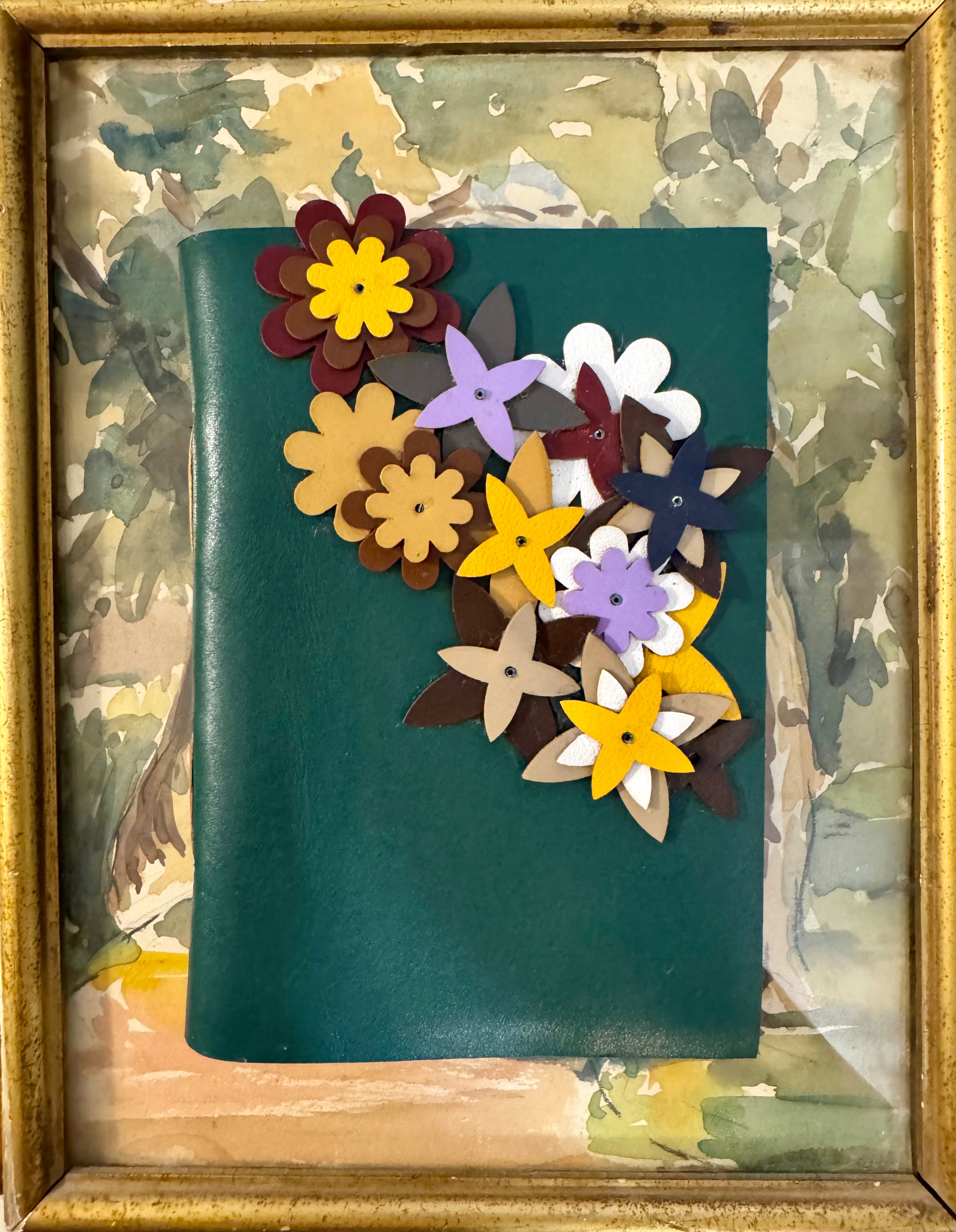 A square dark teal leather journal or notebook cover decorated with layered paper flowers in yellow, purple, brown, white, and navy, displayed inside a gold-toned frame over a watercolor-style background.