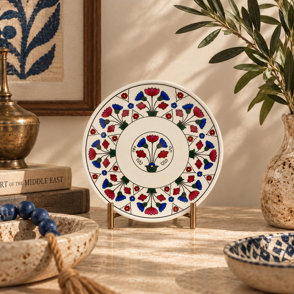 A round white ceramic plate with intricate red and blue floral hand-painted patterns, displayed on a small gold stand on a stone tabletop.